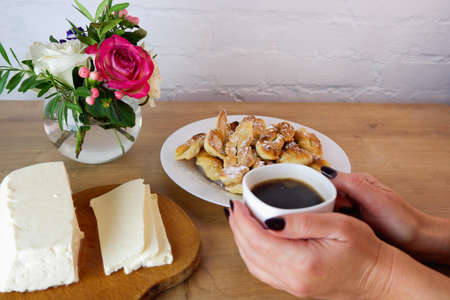 Cup of coffee in female hands, close-up. Homemade baked goods during quarantine. Homemade dessert preparation. Homemade feta cheese on a wooden tabletop. Concept on the background of a white brick wall and a bouquet of roses.の写真素材