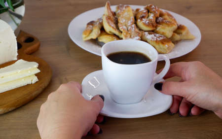 Cup of coffee in female hands, close-up. Homemade baked goods during quarantine. Homemade dessert preparation. Homemade feta cheese on a wooden tabletop. Concept on the background of a white brick wall and a bouquet of roses.の写真素材