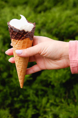 A woman's hand with a manicure holds a chocolate waffle cone of ice cream on the background of a...