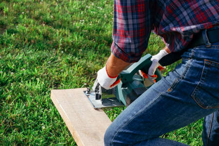 Working with a power tools. A builder is sawing a board at the construction site of a new house. Close-up on gloved hands holding a circular saw.の写真素材