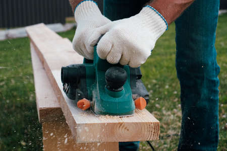 Carpenter hands in gloves electric plane planing lumber, flying shavings, sawdust. Close-up on...