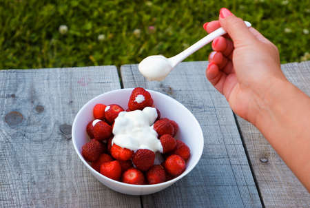 A female hand on a wooden bench eats a strawberry dessert with cream for breakfast.の写真素材