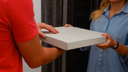Pizza delivery service. A courier in a red polo hands pizza to a young woman at the door of her house.の写真素材