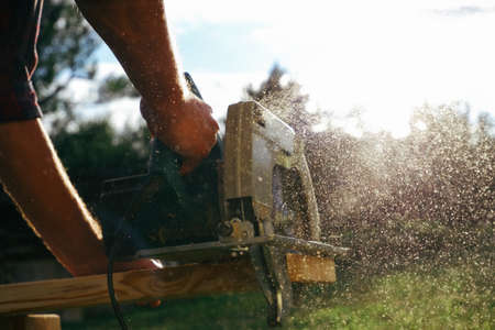 A male carpenter works with a circular saw on the lawn. Close-up view on hands of a working tool. Contrast sunlight with glare scattered in flying sawdust.の写真素材