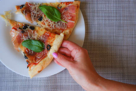 Close-up top view of a female hand takes a slice of pizza from a white plate.の写真素材