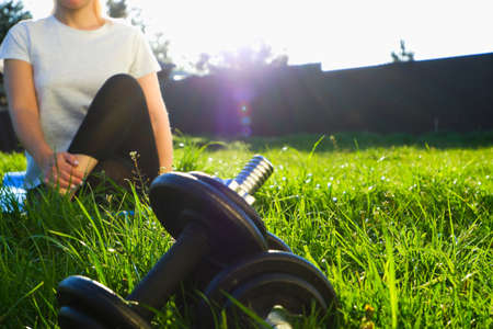 Dumbbells on green grass in the backlight of the setting sun. The woman in the background stretches after a hard workout.の写真素材