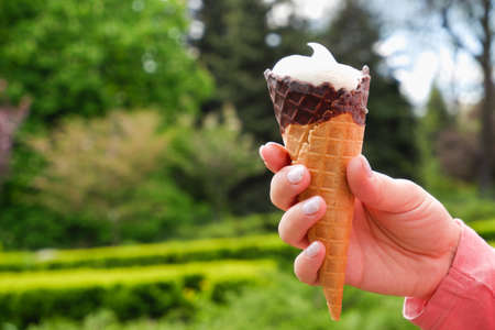 A woman's hand with a manicure holds a chocolate waffle cone of ice cream on the background of a...