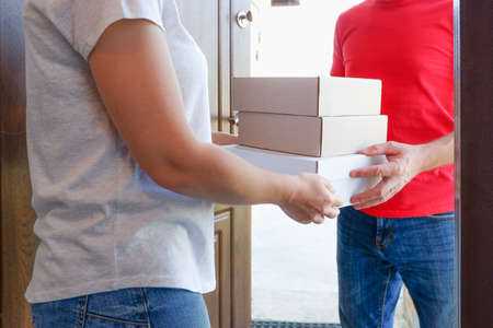 A delivery courier hands over cardboard boxes of goods to a woman near the door of her house.
