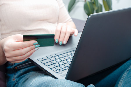 A woman at home sitting on a sofa holds a credit card in her hands and makes a payment using a laptop. happy online shopper. Woman using credit card to purchase merchandise on internetの写真素材