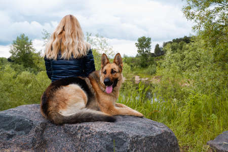A young woman with a German Shepherd sits on a large granite stone by the lake on a cloudy spring day.の写真素材