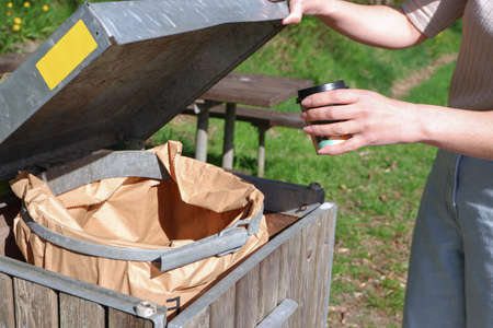 A woman throws a cardboard cup after coffee with a plastic lid into a paper container. Coffee in a...