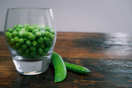 Green peas in glass bowl. fresh pea in the pod with green leaves. green peas on a brown wodden table. Shelled peas in a glass with the pods on the sideの写真素材