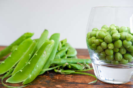 Green peas in glass bowl. fresh pea in the pod with green leaves. green peas on a brown wodden table. Shelled peas in a glass with the pods on the sideの写真素材