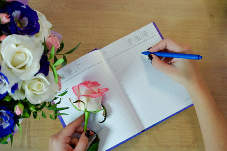 Female hands writing in open notebook and bouquet of roses on old wooden table. Planning notebook with pen, woman makes entry in her diary on table.の写真素材
