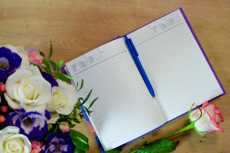 Notepad, diary with a pen on a wooden table near a bouquet of multi-colored roses. Workspace with...