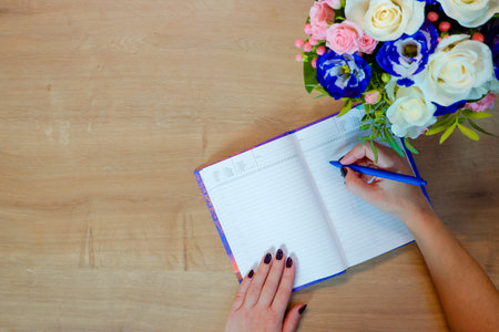 Female hands writing in open notebook and bouquet of roses on old wooden table. top view. Hand writing a love note in a notebook.の写真素材