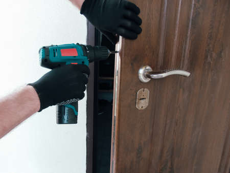The master worker installs a door lock in the front door, metal doors with a polymer coating, using an electric drill screwdriver, close-up.の写真素材
