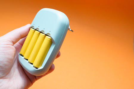 Charger and Batteries in male hand. Male hand holding charger with yellow AA batteries, on on orange...