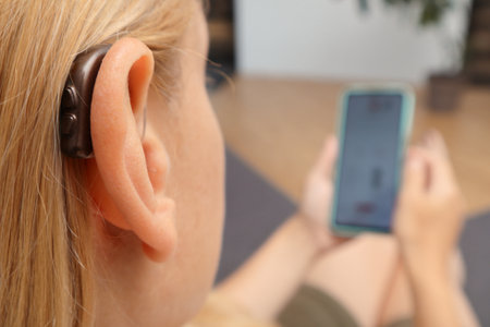 Young Hearing woman impaired using smartphone indoors. Close up of blonde woman wearing hearing aid on ear and pressing button while using smartphone at home.の写真素材