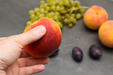 Ripe delicious peach in hand with organic fruit in the background. Plums, grapes, peaches.の写真素材