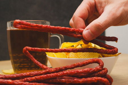 A man's hand takes snacks next to beer. Beer snack. Dry thin meat sausages and chips. Man drinking beer and eating snacksの写真素材