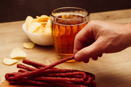 A man's hand takes snacks next to beer. Beer snack. Dry thin meat sausages and chips. Man drinking beer and eating snacksの写真素材