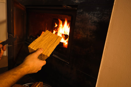 Man putting wood into a fire place. Hand with a log near the open fireplace door. Renewable energy sources.の写真素材