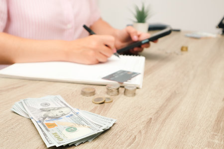 A female shopper holds a mobile phone in her hands and banknotes and credit card next to her. Shopping online using smartphone. Consumerism and financial savingsの写真素材