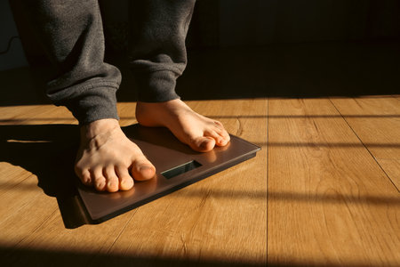 Male is checking her weight on the scale. Feet on a scale for weight control after bathroom. Man standing on weight scale at home. Sun shadows.の写真素材