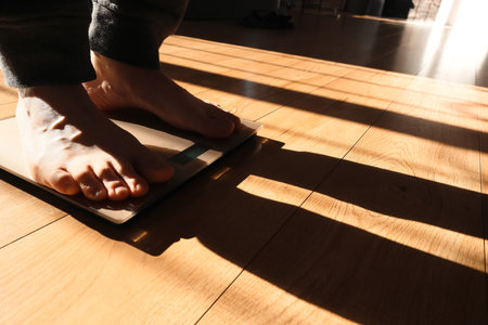 Person is checking their weight on the scale. Feet on a scale for weight control after bathroom. Person standing on weight scale at home. Sun shadows.の写真素材