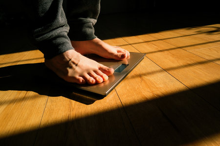 Person checking their weight on the scale. Feet on a scale for weight control after bathroom. Person standing on weight scale at home. Sun shadows.の写真素材