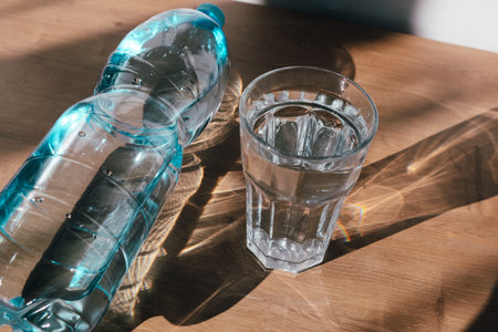 Water blue plastic bottle in glass, with sunlight deep shadow of glass. Hard sunlight. Water bottle of transparent plastic filling a drinking glass.の写真素材