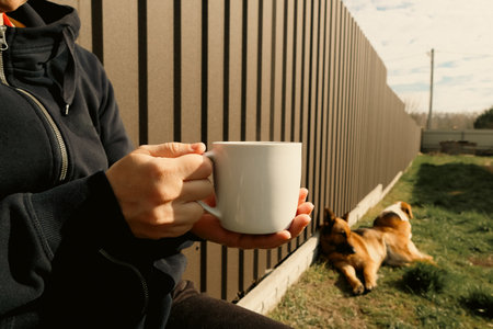 The first cup of coffee on a sunny morning in the backyard with a dog. A woman's hand holds a cup with a hot drink. Woman enjoying coffee in garden with dogの写真素材