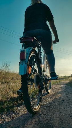 Rear view of a person riding a bike along a countryside dirt path in the evening sunlight. Active...