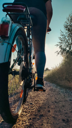 Rear view of a person riding a bike along a countryside dirt path in the evening sunlight. Active...