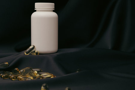 Minimalistic photo of a white plastic pill bottle with scattered yellow capsules on a dark fabric background. Perfect for concepts related to health, medicine, pharmacy, vitamins, supplements, nutrition, and wellness advertising.の写真素材