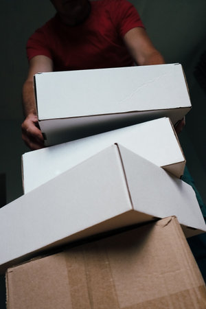 Low-angle view of a man in a red t-shirt holding and lifting a stack of white cardboard boxes. Concept of moving, relocation, delivery, packaging, or logistics in a new home or office.の写真素材