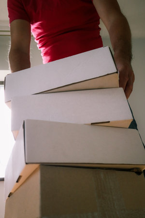 Low-angle view of a man in a red t-shirt holding and lifting a stack of white cardboard boxes....