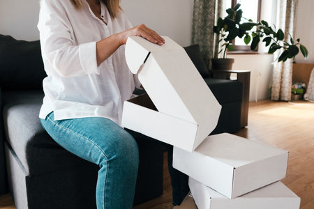 Close-up of a woman sitting on a sofa and unpacking white cardboard boxes in a bright living room....
