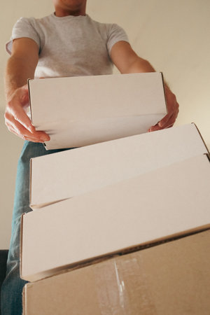 Low-angle view of a man in a t-shirt holding and lifting a stack of white cardboard boxes. Concept of moving, relocation, delivery, packaging, or logistics in a new home or office.の写真素材