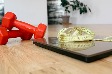 Close-up of red dumbbells, a measuring tape, and a digital scale on a wooden floor. Concept of...