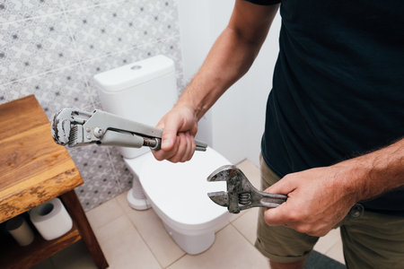 A man, a plumber, is holding two used adjustable wrenches in his hands while standing in a bathroom with a white toilet and patterned tiles in the background. Plumbing repair service.の写真素材