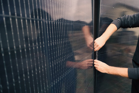 Close-up of a person's hands plugging together male and female MC4 connectors. The black cables are being connected to power a solar energy system installed on a residential fenceの写真素材