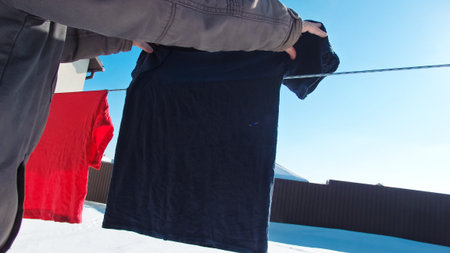 A person hangs a dark t-shirt on a wire line against a blue sky. The backyard is covered in snow...