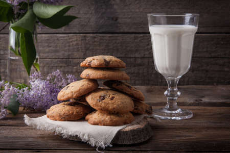 Stack of freshly baked chocolate chip cookies with glass of milk lying on linen napkin on rustic wooden tableの写真素材