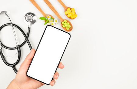 Female hand with blank screen smartphone with stethoscope and organic pills in wooden spoons in the backdrop. Telemedicine conceptの写真素材