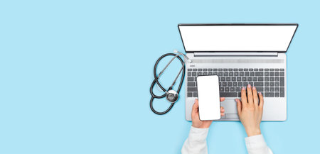 Female doctor's hands holding smartphone with blank screen and using laptop with empty monitor on blue background. Top view with copy space, stethoscope near the laptop. Telemedicine conceptの写真素材