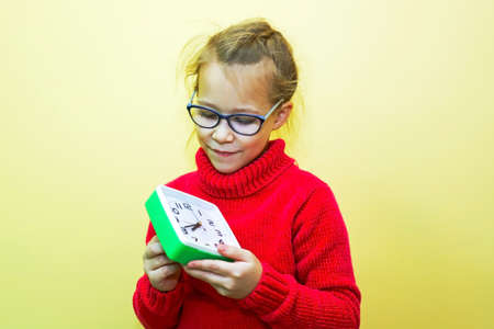 A child girl in a red sweater plays with an alarm clock, looks at the time and makes a gesture,...