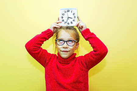 A child girl in a red sweater plays with an alarm clock, looks at the time and makes a gesture, showing the time or being lateの写真素材
