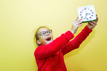 A child girl in a red sweater plays with an alarm clock, looks at the time and makes a gesture, showing the time or being lateの写真素材